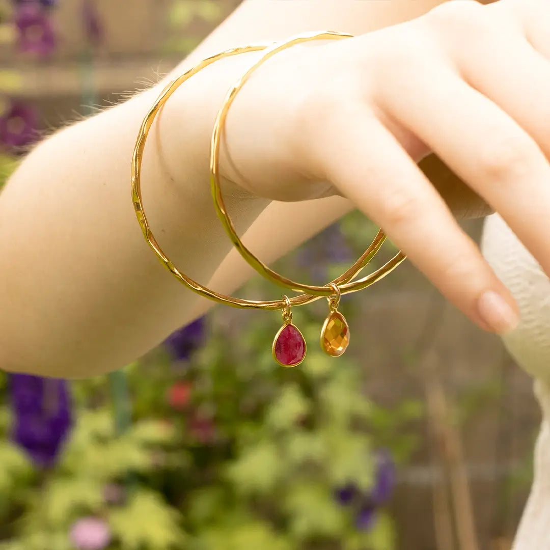 Two gold hammered bangles with ruby and citrine teardrop gemstone charms, worn on wrist in soft natural light with blurred garden background