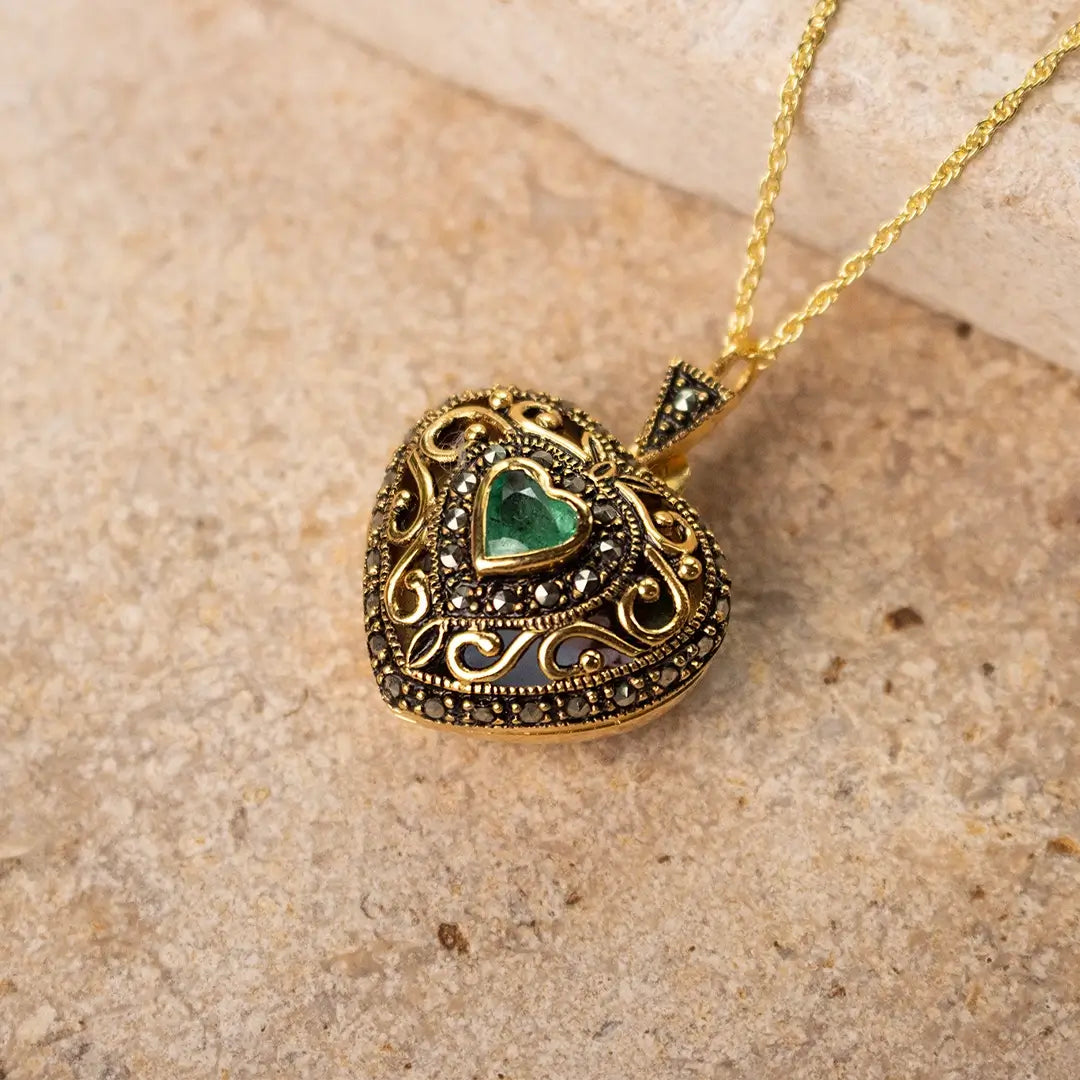 A close-up of a gold heart-shaped locket with intricate detailing and a green heart-shaped emerald gemstone at its center, lying on a textured beige stone surface.