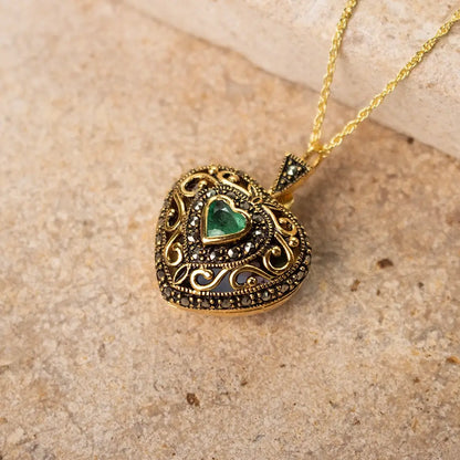 A close-up of a gold heart-shaped locket with intricate detailing and a green heart-shaped emerald gemstone at its center, lying on a textured beige stone surface.