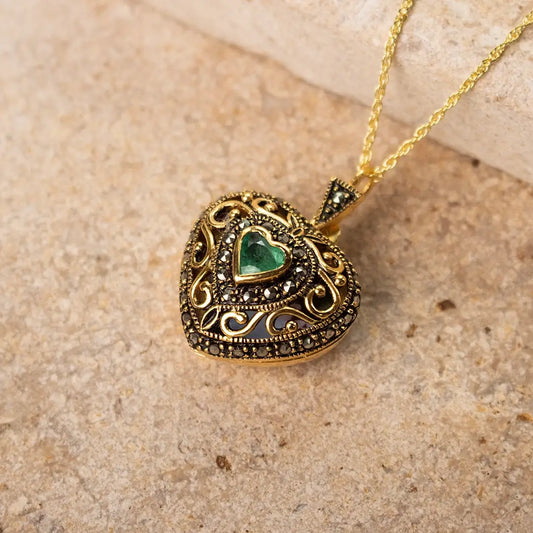 A close-up of a gold heart-shaped locket with intricate detailing and a green heart-shaped emerald gemstone at its center, lying on a textured beige stone surface.