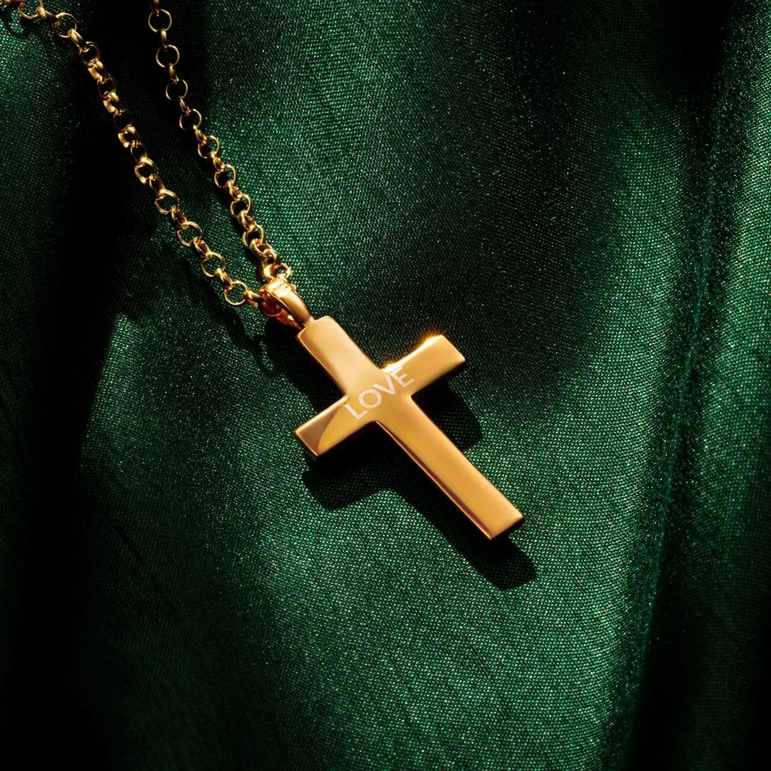A gold heavy cross pendant by Lily Blanche engraved with the word “LOVE,” hanging from a gold chain and displayed against a deep green fabric backdrop with soft, dramatic lighting.
