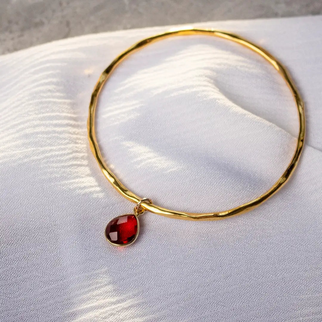 Gold hammered bangle with single garnet teardrop charm resting on white fabric, highlighting the rich red gemstone and polished finish