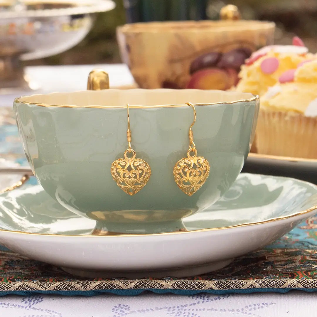 Gold filigree heart drop earrings displayed on a green teacup and saucer, styled with a vintage tea setting including cupcakes and fruit in the background