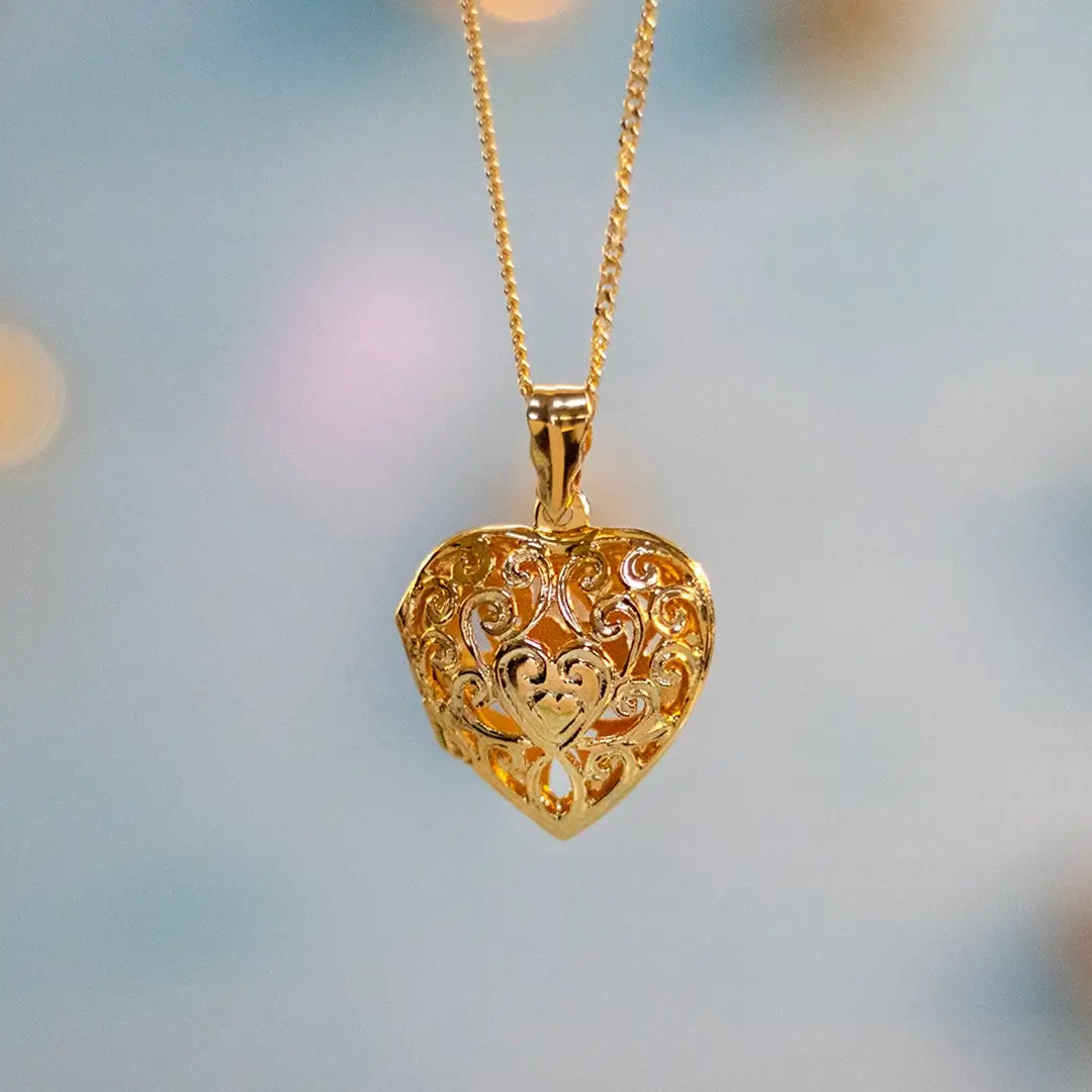 A gold heart-shaped locket pendant with an intricate filigree design, hanging from a delicate gold chain against a softly blurred background with warm light.