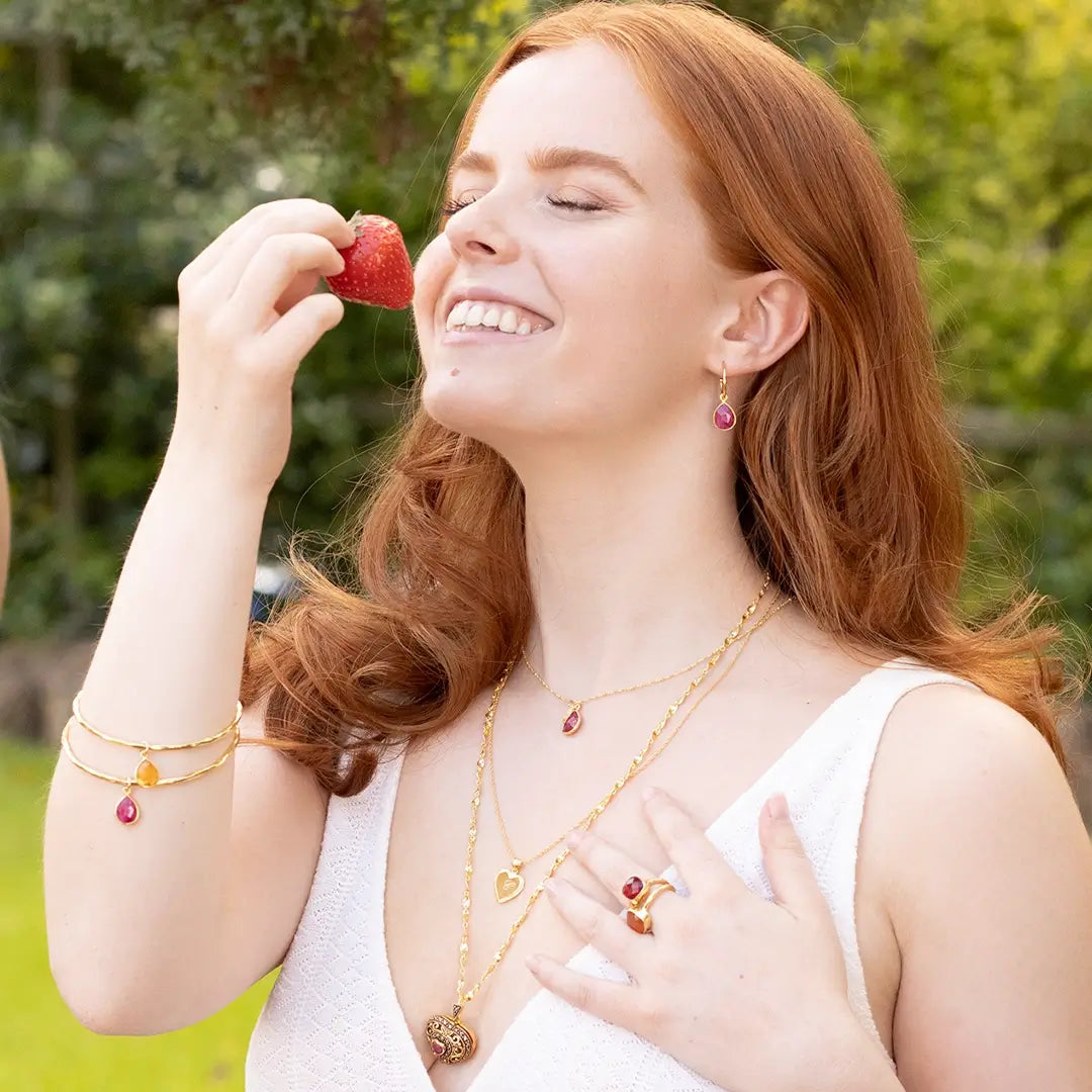 Woman wearing layered gold jewellery including gemstone necklaces, rings, earrings and stacked bangles with ruby and citrine charms, holding a strawberry outdoors in sunlight
