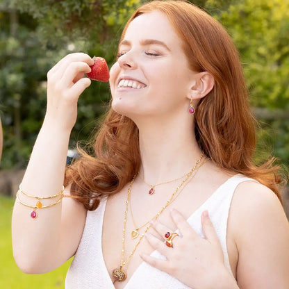 Woman wearing layered gold jewellery including gemstone necklaces, rings, earrings and stacked bangles with ruby and citrine charms, holding a strawberry outdoors in sunlight