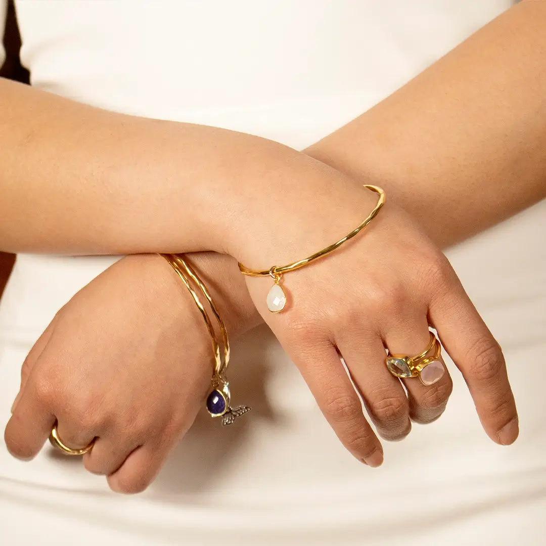 Gold hammered bangles with moonstone and lapis lazuli teardrop charms, worn on wrists alongside gold gemstone rings, styled against a soft neutral background