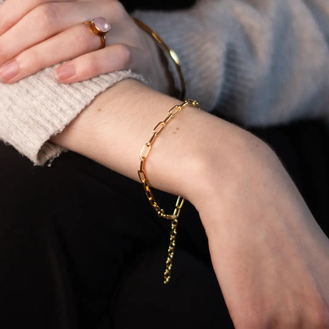 A close-up shot of a fair-skinned arm wearing a chunky gold paperclip chain bracelet and a thin gold bangle. The hand has a gold ring with a soft pink stone on the ring finger. The person is wearing a light beige, cozy-looking sweater and dark trousers, sitting in soft, natural light indoors or outside.