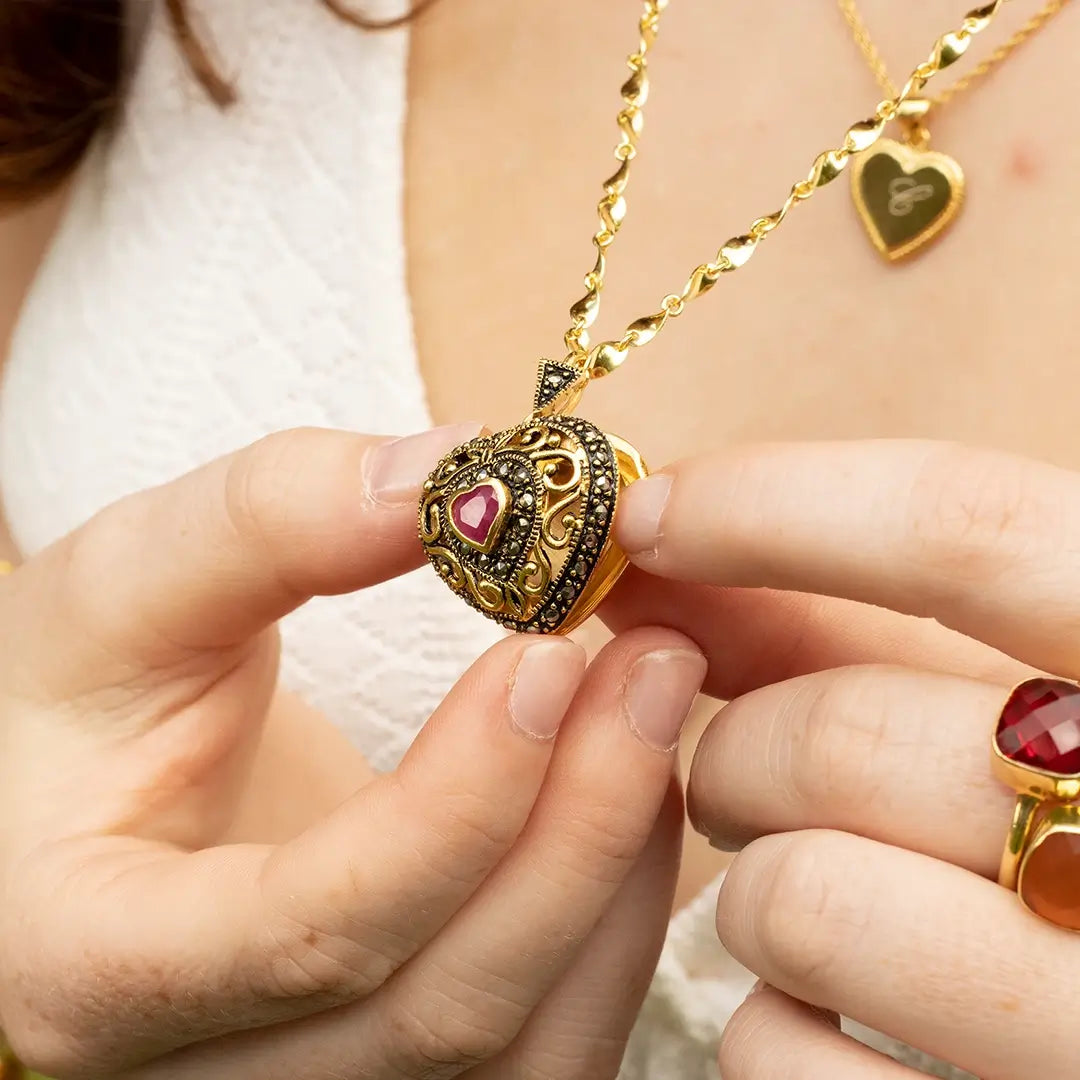 Close-up of a vintage-style gold heart locket with ornate detailing and a central ruby gemstone, held by hand on a gold chain, with a matching ruby ring visible.