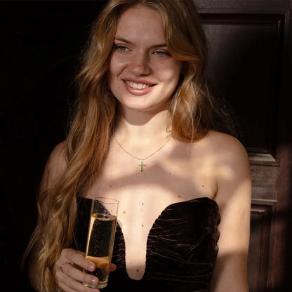 A woman with long light brown hair smiles while holding a champagne glass. She’s wearing a strapless dark velvet dress and a gold necklace featuring a tsavorite cross pendant. Warm sunlight highlights her face and neckline against a dark wooden background.