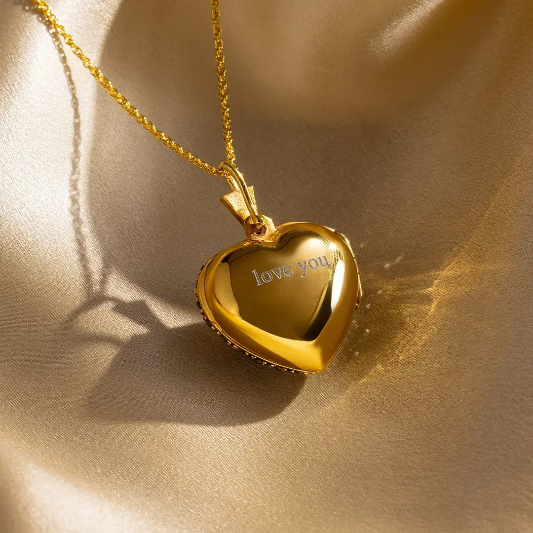 Gold heart locket necklace engraved with “love you” in a classic font, displayed on soft gold satin with warm lighting.