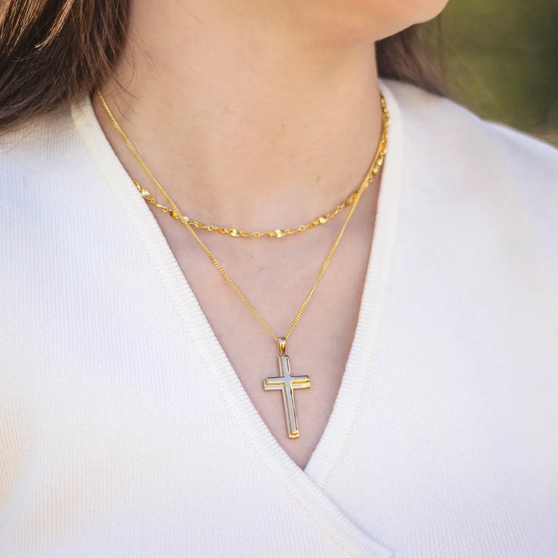 woman wearing a white v neck top with two layered gold necklaces. The shorter necklace is a decorative twist chain and the longer necklace is a simple chain with a large gold cross pendant