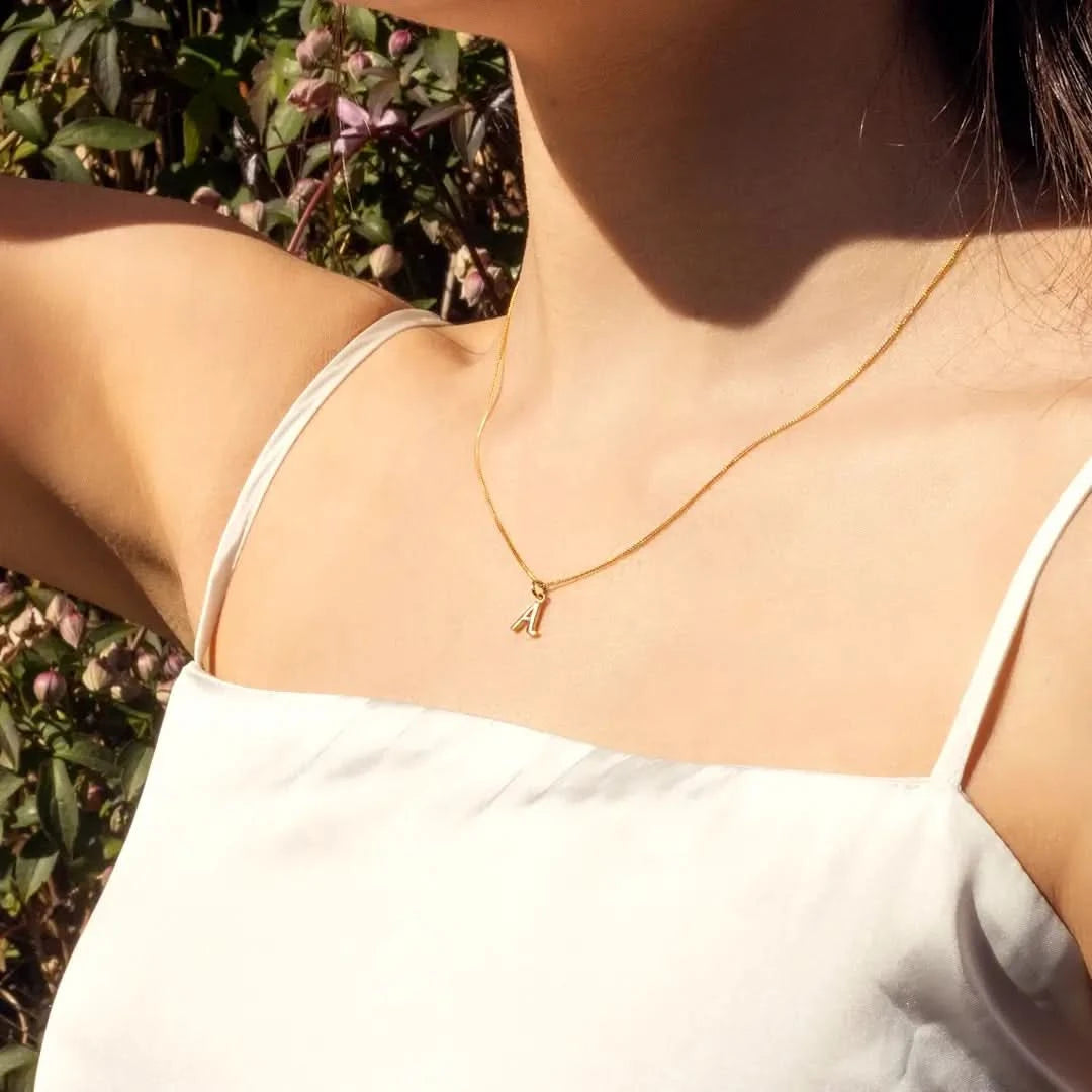 Close-up of a female model wearing a white strappy top and the 9-carat solid gold initial necklace, letter 'A', on a close curb chain against a background of greenery.