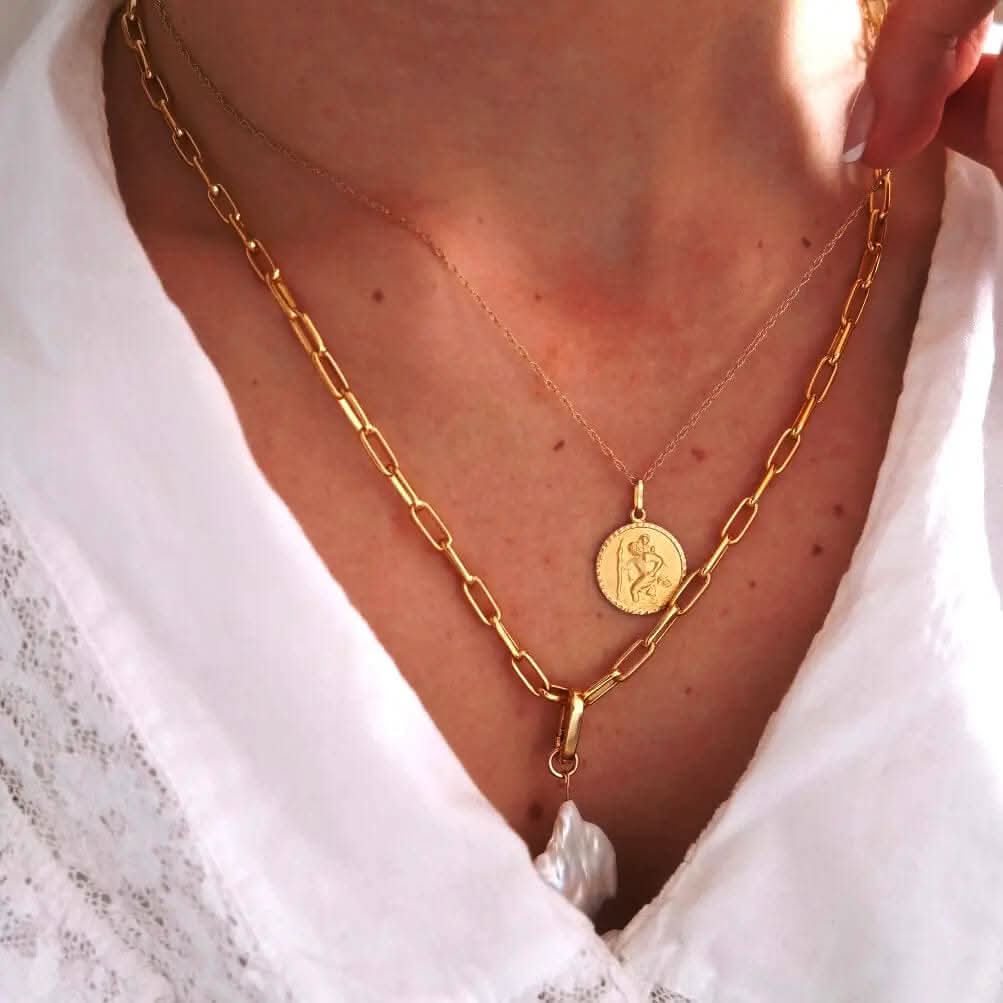 Close-up image of a woman wearing layered gold necklaces, including a fine chain with a gold St. Christopher pendant and a chunky paperclip chain with a pearl drop