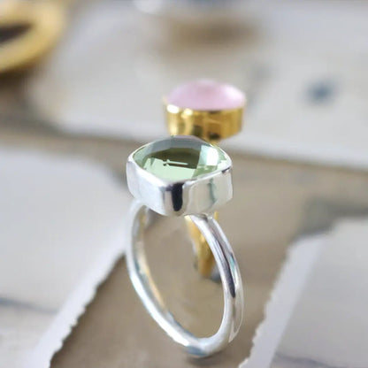 Close-up of a sterling silver ring with a faceted green amethyst gemstone, with a gold ring featuring a pink stone softly blurred in the background.