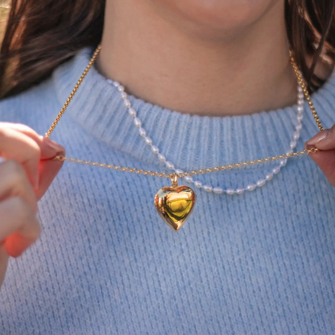 Model holding gold heart locket necklace on belcher chain over light blue sweater