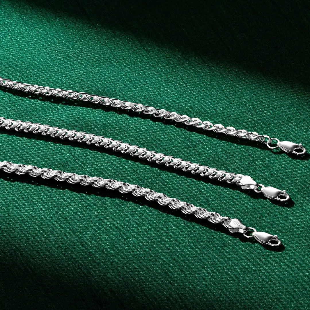Three silver chain necklaces laid out horizontally on a deep green fabric background. Each chain features a different link style and thickness, with lobster-claw clasps visible on the right side. Dramatic lighting creates shadows and highlights on the metal links.