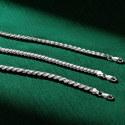 Three silver chain necklaces laid out horizontally on a deep green fabric background. Each chain features a different link style and thickness, with lobster-claw clasps visible on the right side. Dramatic lighting creates shadows and highlights on the metal links.
