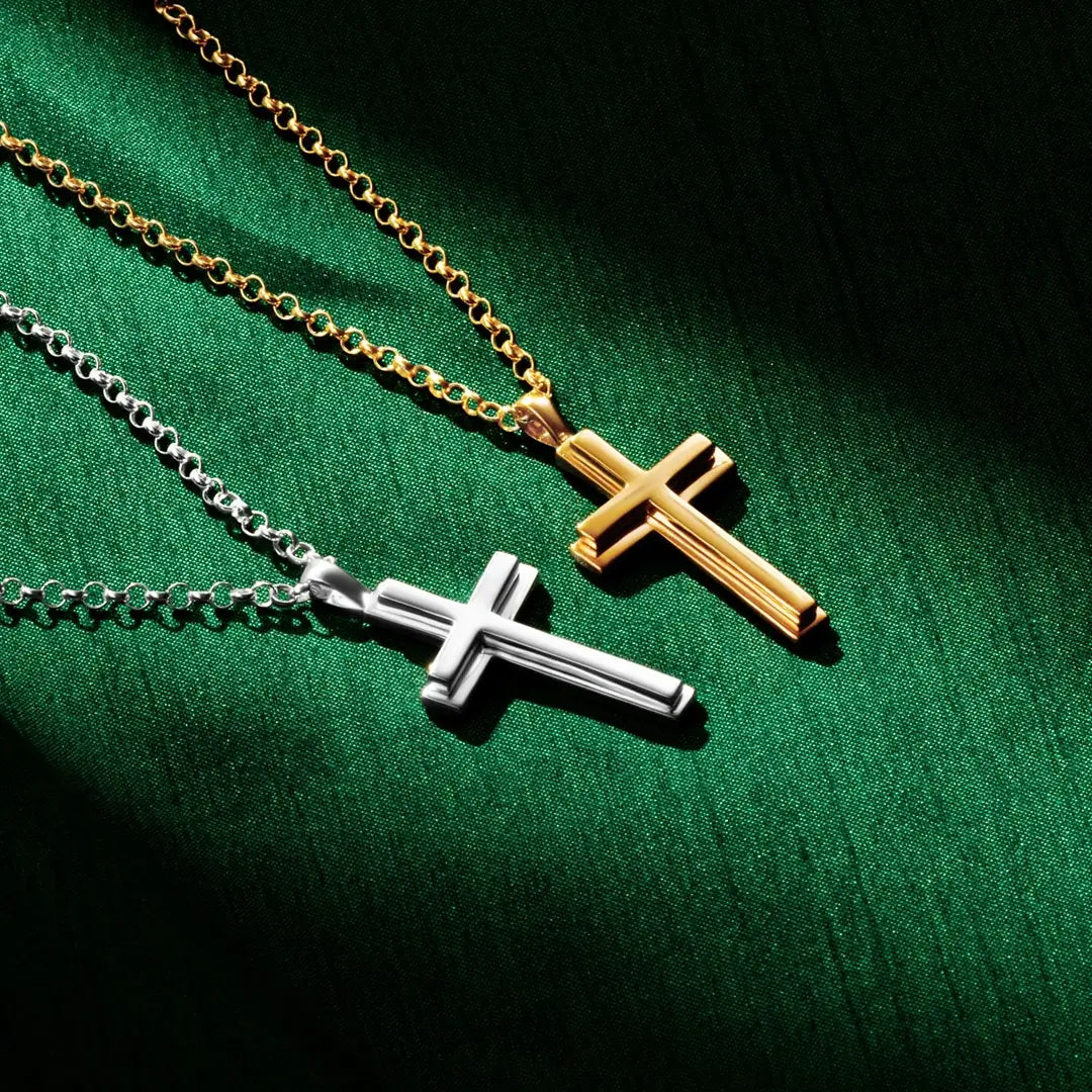 Two cross necklaces, one gold and one silver, displayed on a rich green fabric background. The gold cross hangs from a gold chain in the upper right, while the silver cross hangs from a silver chain below. Both pendants have a polished, beveled design with dramatic lighting creating shadows on the fabric.