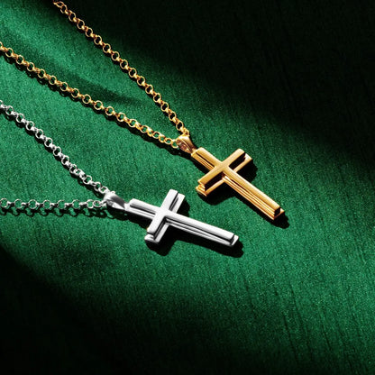 Two cross necklaces, one gold and one silver, displayed on a rich green fabric background. The gold cross hangs from a gold chain in the upper right, while the silver cross hangs from a silver chain below. Both pendants have a polished, beveled design with dramatic lighting creating shadows on the fabric.