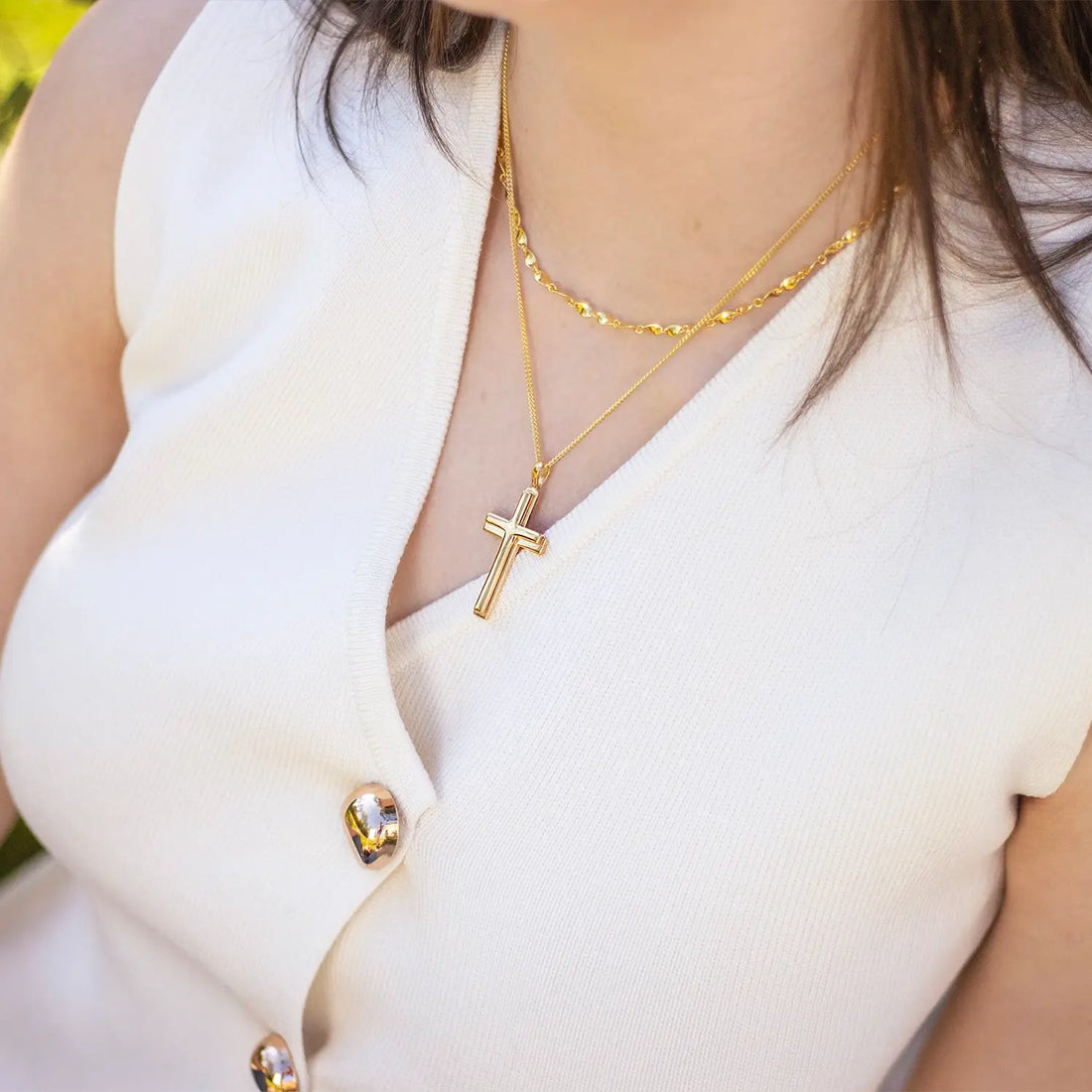 A woman wearing a white sleeveless top with gold buttons, layered with two gold necklaces — one with small heart-shaped links and the other featuring a bold gold cross pendant.