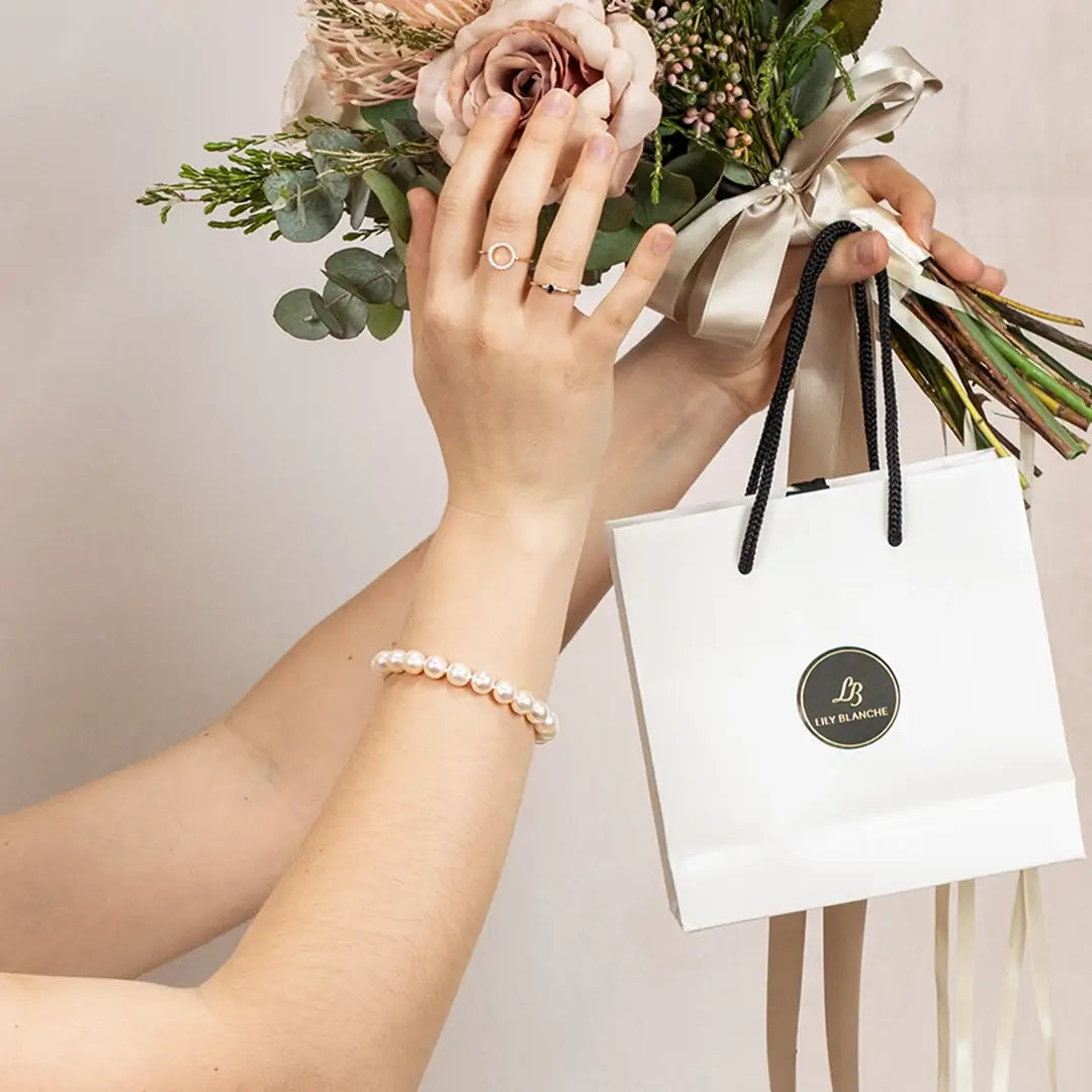 Ivory freshwater pearl bracelet worn on wrist while holding a bouquet of flowers and a Lily Blanche gift bag, highlighting elegant bridal or special occasion styling.