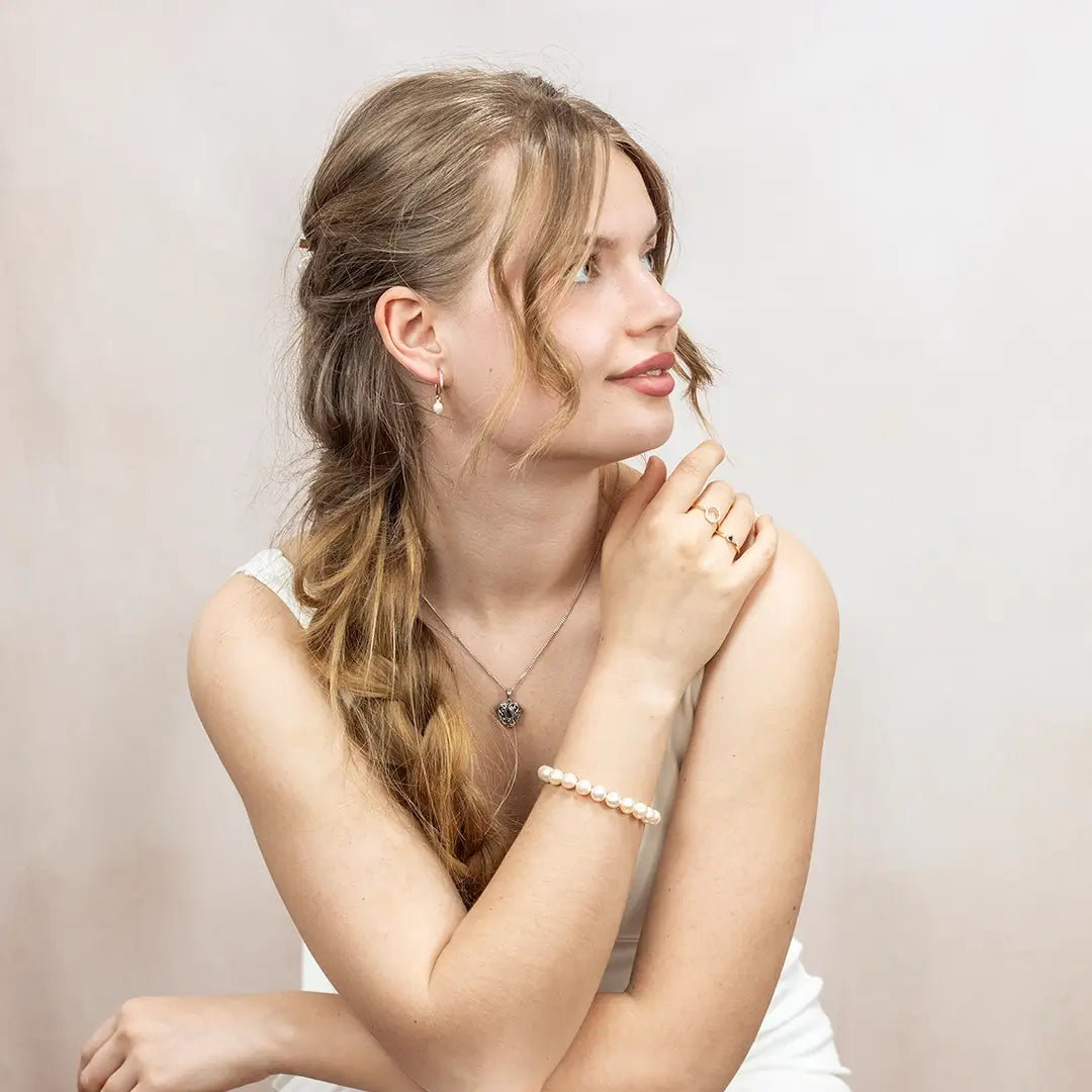 Woman wearing an ivory freshwater pearl bracelet styled with silver jewellery, including drop pearl earrings and a silver heart locket necklace, photographed against a light studio backdrop.