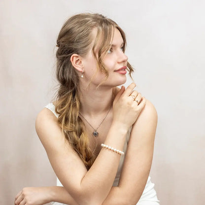 Woman wearing an ivory freshwater pearl bracelet styled with silver jewellery, including drop pearl earrings and a silver heart locket necklace, photographed against a light studio backdrop.
