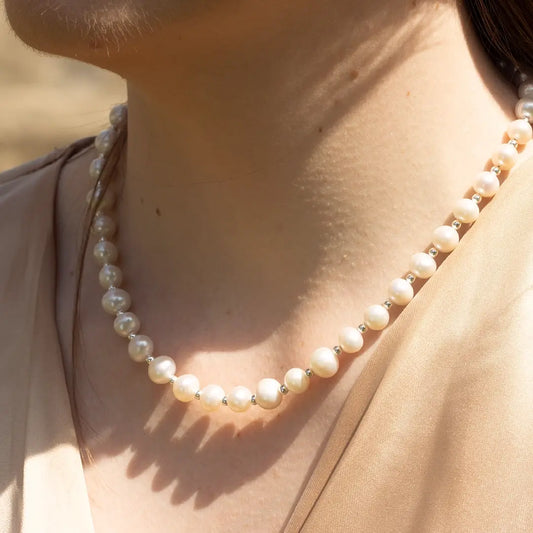 Close-up of an ivory pearl necklace with small silver spacer beads, resting along the neckline and catching natural sunlight against a soft neutral blouse.