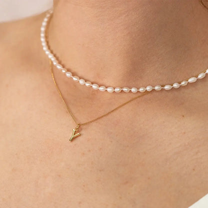 Close-up of ivory freshwater seed pearl choker necklace layered with a fine gold initial pendant, highlighting small oval pearls and delicate gold chain detail.