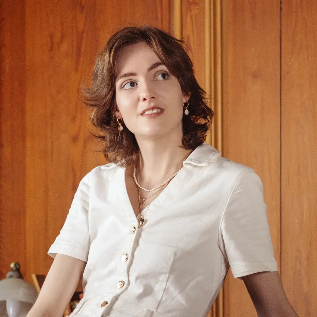 Woman wearing an ivory seed pearl choker necklace paired with gold drop pearl earrings and layered gold pendants, styled with a white blouse against a warm wooden interior background.