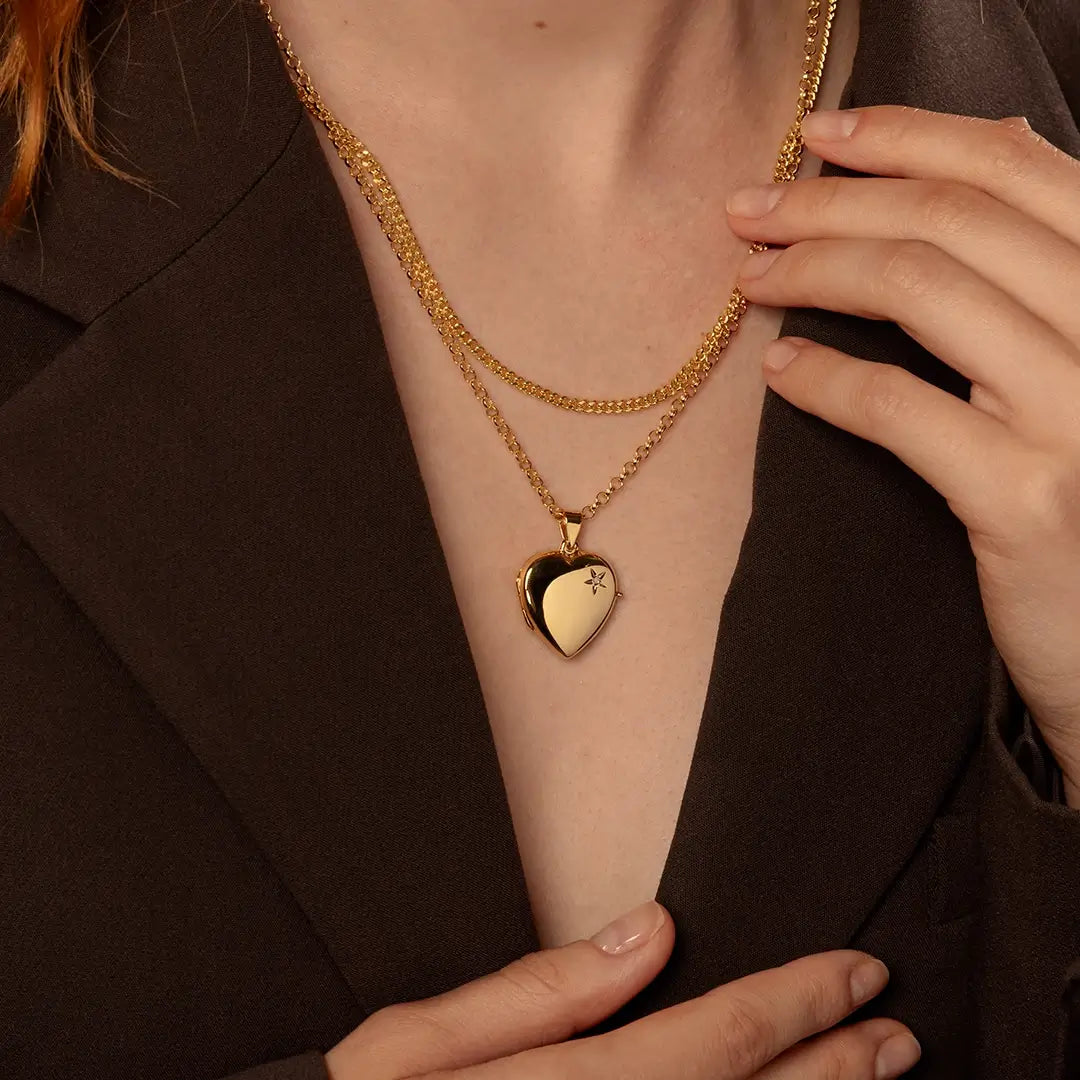 A close-up of a person wearing a thick curb chain layered with a heart-shaped locket pendant on a belcher chain, styled with a dark brown blazer.