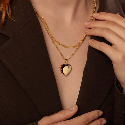 A close-up of a person wearing a thick curb chain layered with a heart-shaped locket pendant on a belcher chain, styled with a dark brown blazer.