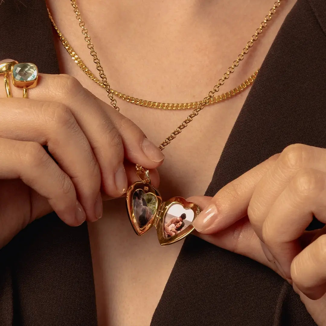 Close-up of a person wearing layered gold necklaces, including a heart-shaped locket pendant with two photos inside. The person is wearing a dark brown top and a gold ring on one hand.