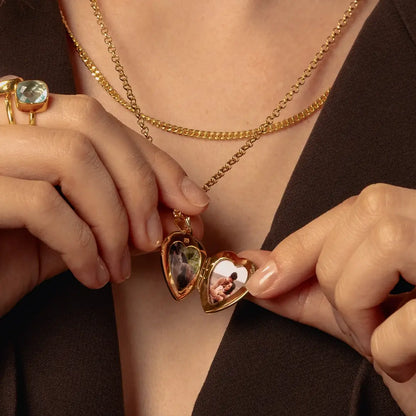 Close-up of a person wearing layered gold necklaces, including a heart-shaped locket pendant with two photos inside. The person is wearing a dark brown top and a gold ring on one hand.