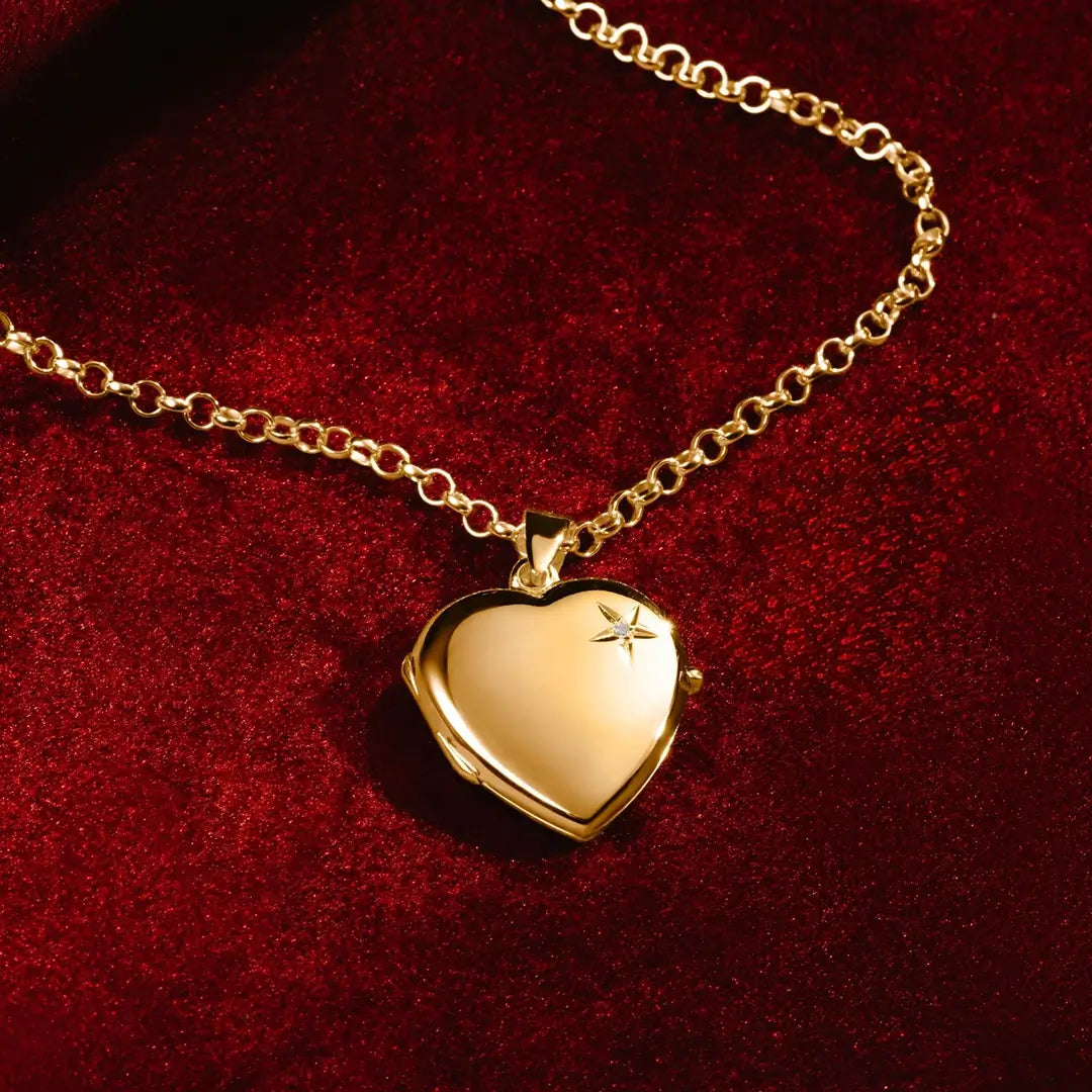 A gold heart-shaped locket with a small diamond on the front, displayed on a rich red velvet background. The locket hangs from a gold chain, catching the light with a polished shine.