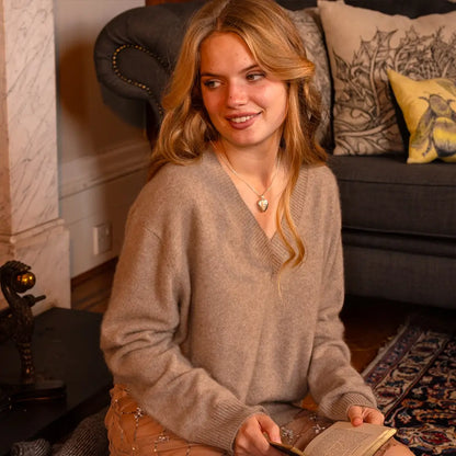 A woman wearing a beige sweater and soft waves in her hair smiles while holding a book, accessorized with a silver heart locket necklace.