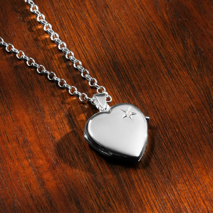 A silver heart-shaped locket with a small diamond rests on a warm wooden surface. The silver chain is arranged above the locket, which shines with a smooth, polished finish.