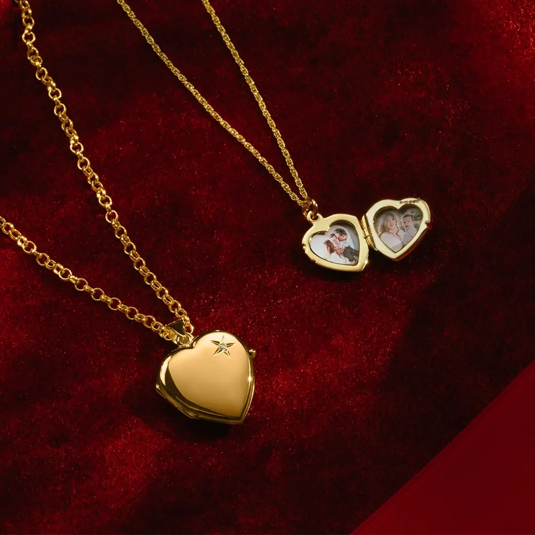 Two gold heart lockets displayed on red velvet: one closed with a diamond on the front, the other open to reveal small photo inserts. Both are shown with their gold chains arranged decoratively.