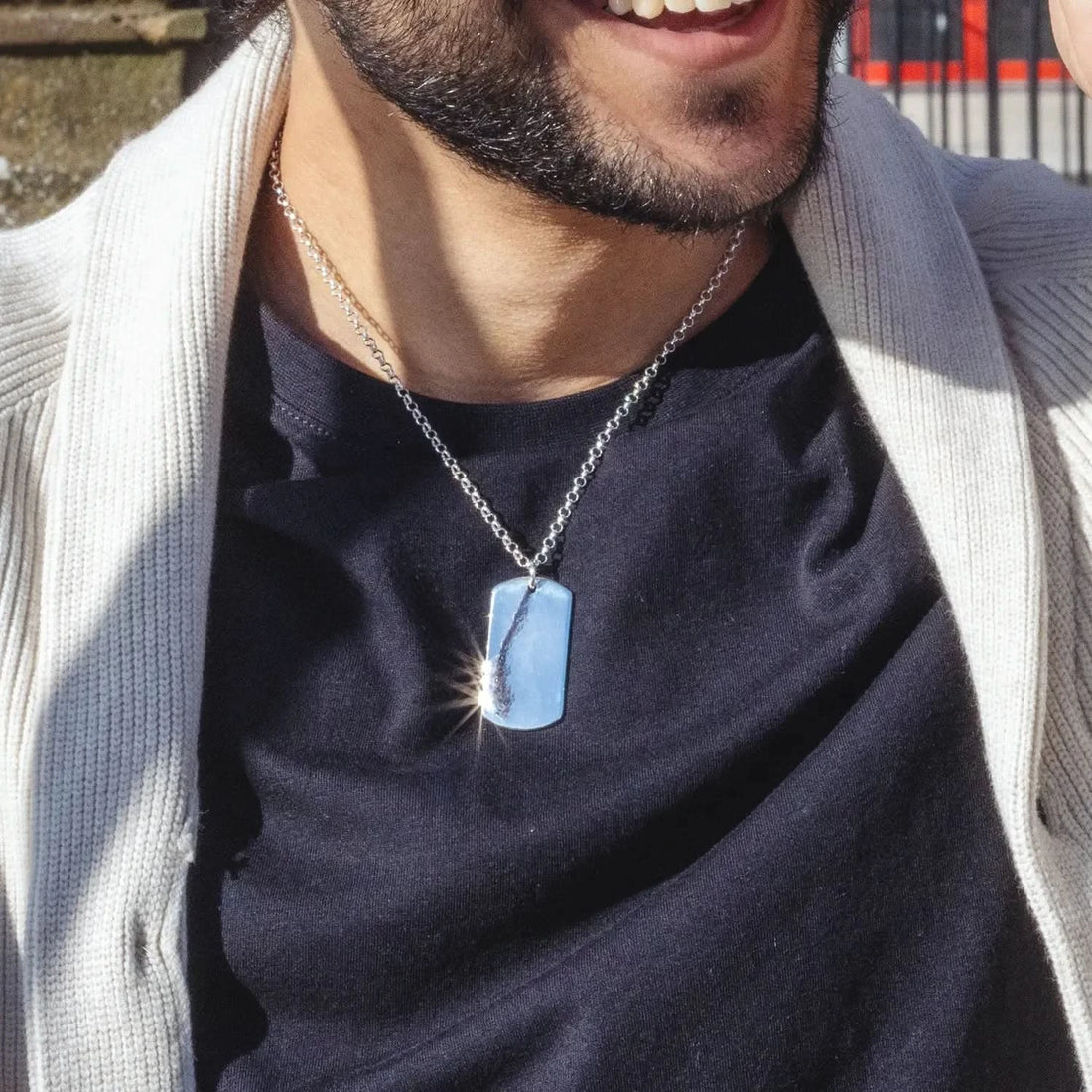 Solid silver extra-large dog tag worn by a male modelw wearing a black top and white knitted jacket. The sun is catching the necklace and making it sparkle.