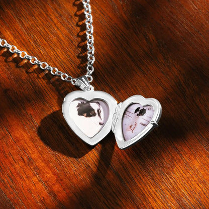 An open silver heart-shaped locket on a wooden surface, revealing two small inserted photos of a black-and-white dog. The silver chain lies loosely above the locket, and the warm wood grain creates a contrasting background.