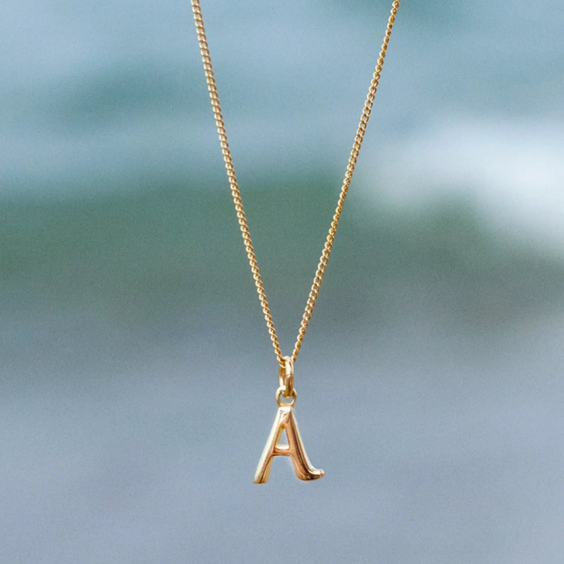 photograph of a  9-carat solid gold initial necklace, letter 'A', on a close curb chain, against a blue sea backdrop