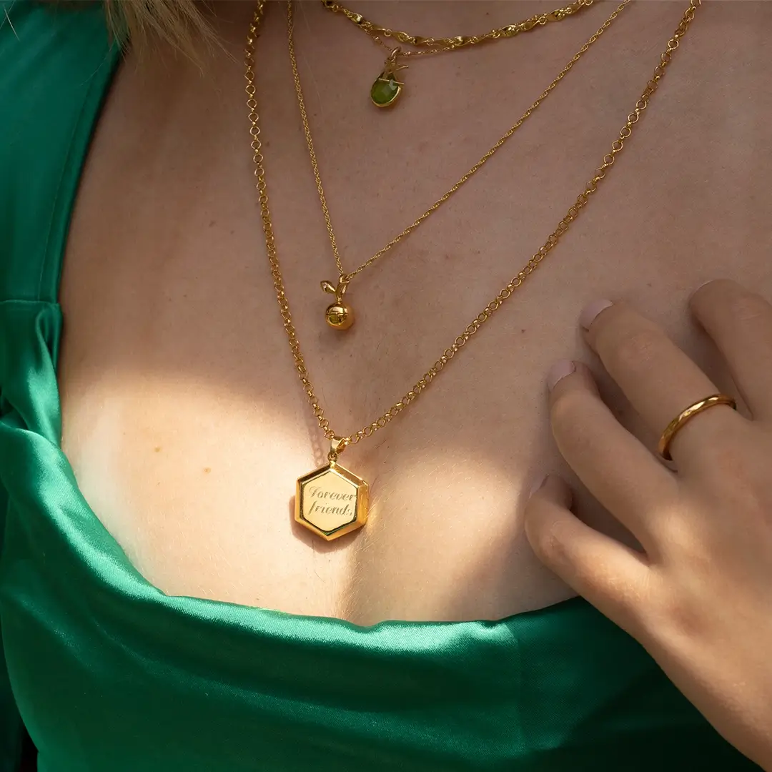 Close-up of layered gold necklaces including an apple charm pendant and a hexagon locket engraved with “Forever Friends”, worn with a green satin dress.