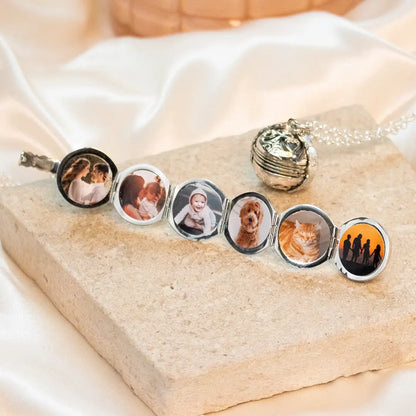 Silver Memory Keeper locket unfolded to display six family and pet photographs beside a closed locket on pale stone.