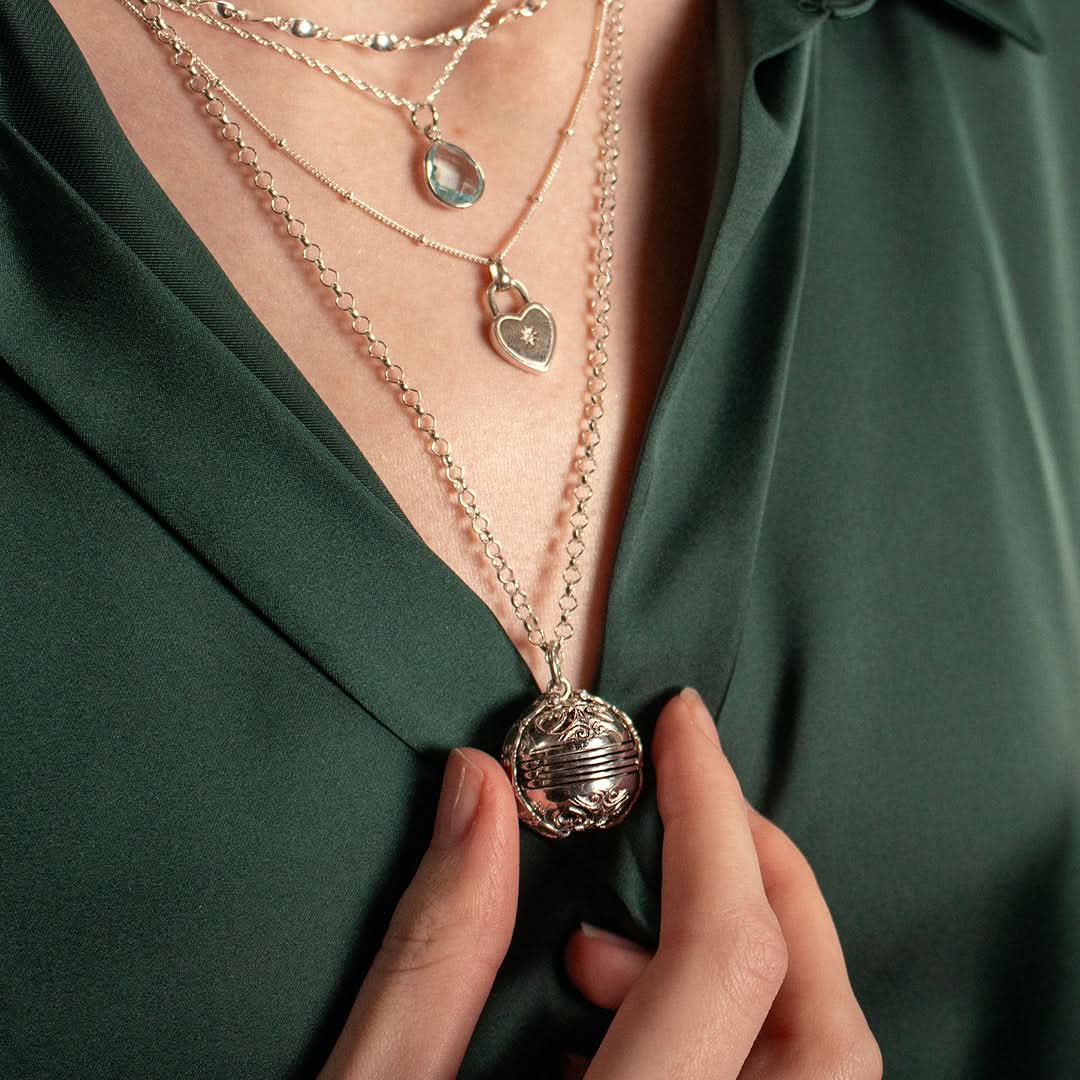 Model's in a green shirt wearing layerd silver necklaces and holding the sterling silver Memory Keeper Locket between thumb and forefinger.