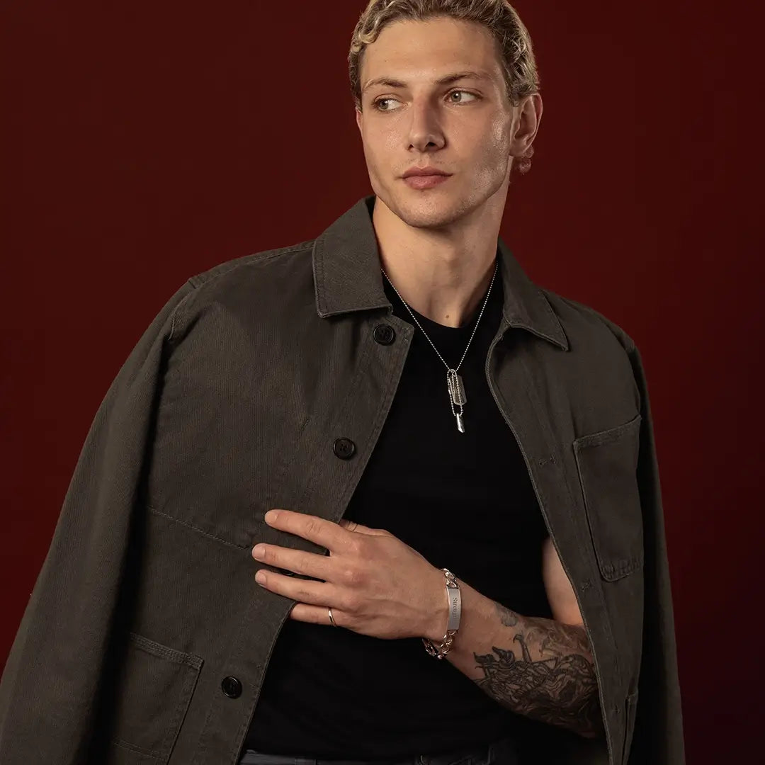 Male model wearing a sterling silver double dog tag necklace and engraved silver ID bracelet against a dark red background