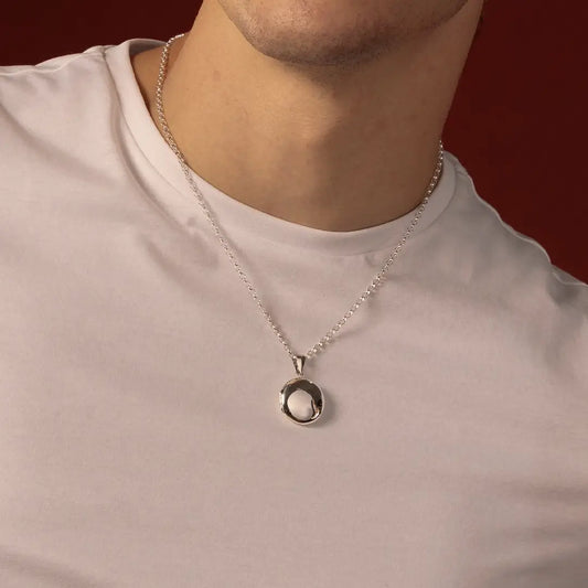 A close-up of a man wearing a silver necklace with a polished round locket, styled with a light-colored t-shirt against a dark red background.