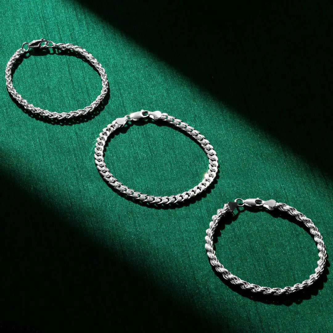 Three silver chain bracelets arranged on a rich green fabric backdrop. Each bracelet has a distinct chain style—from braided to curb to rope—and all are fastened with silver lobster-claw clasps. A diagonal shadow adds contrast to the composition.