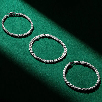 Three silver chain bracelets arranged on a rich green fabric backdrop. Each bracelet has a distinct chain style—from braided to curb to rope—and all are fastened with silver lobster-claw clasps. A diagonal shadow adds contrast to the composition.
