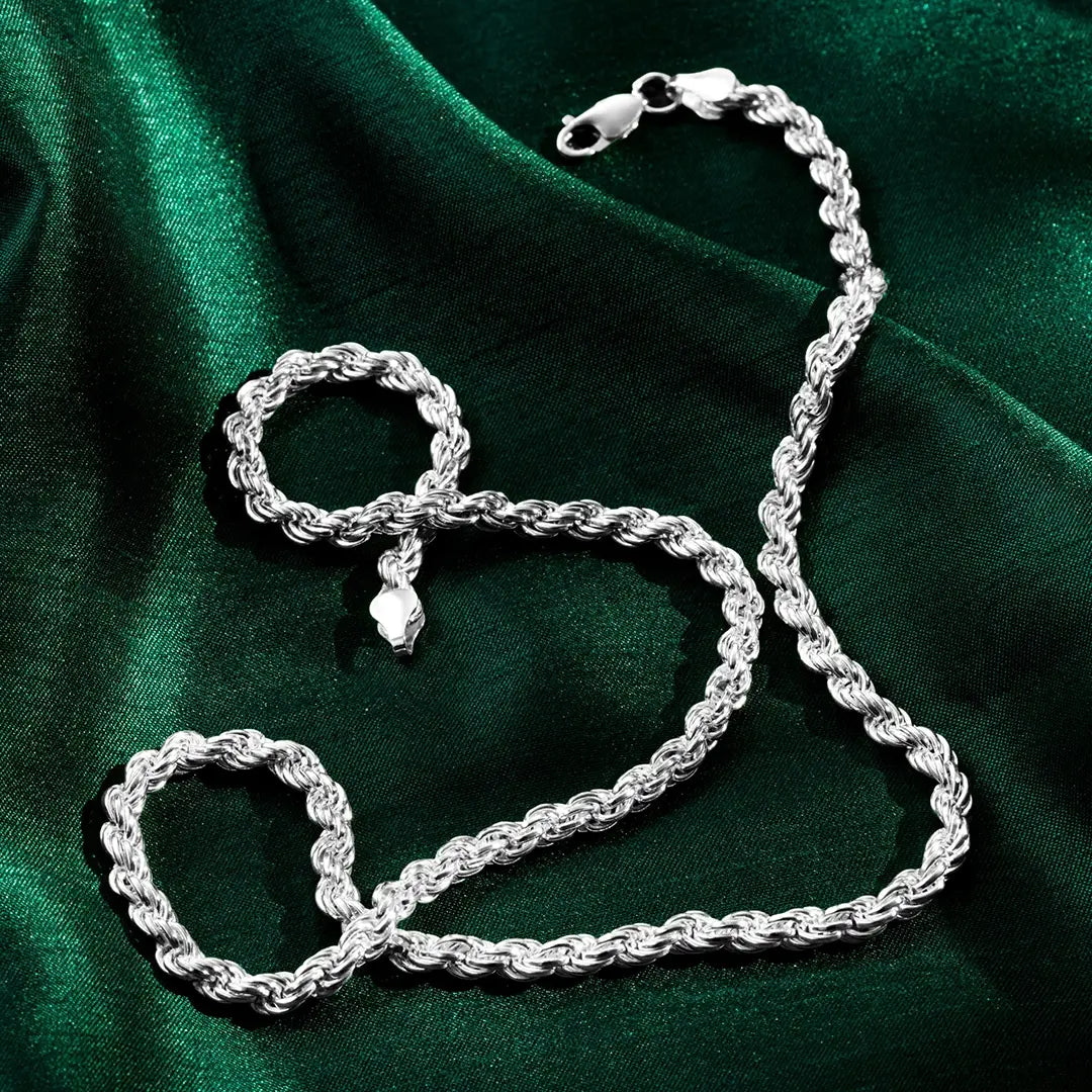 A silver rope-style chain and matching bracelet displayed on a deep green satin fabric. The twisted links create a textured, reflective surface, and both pieces have lobster-claw clasps. Soft highlights and shadows emphasize the draped fabric and the shine of the metal.